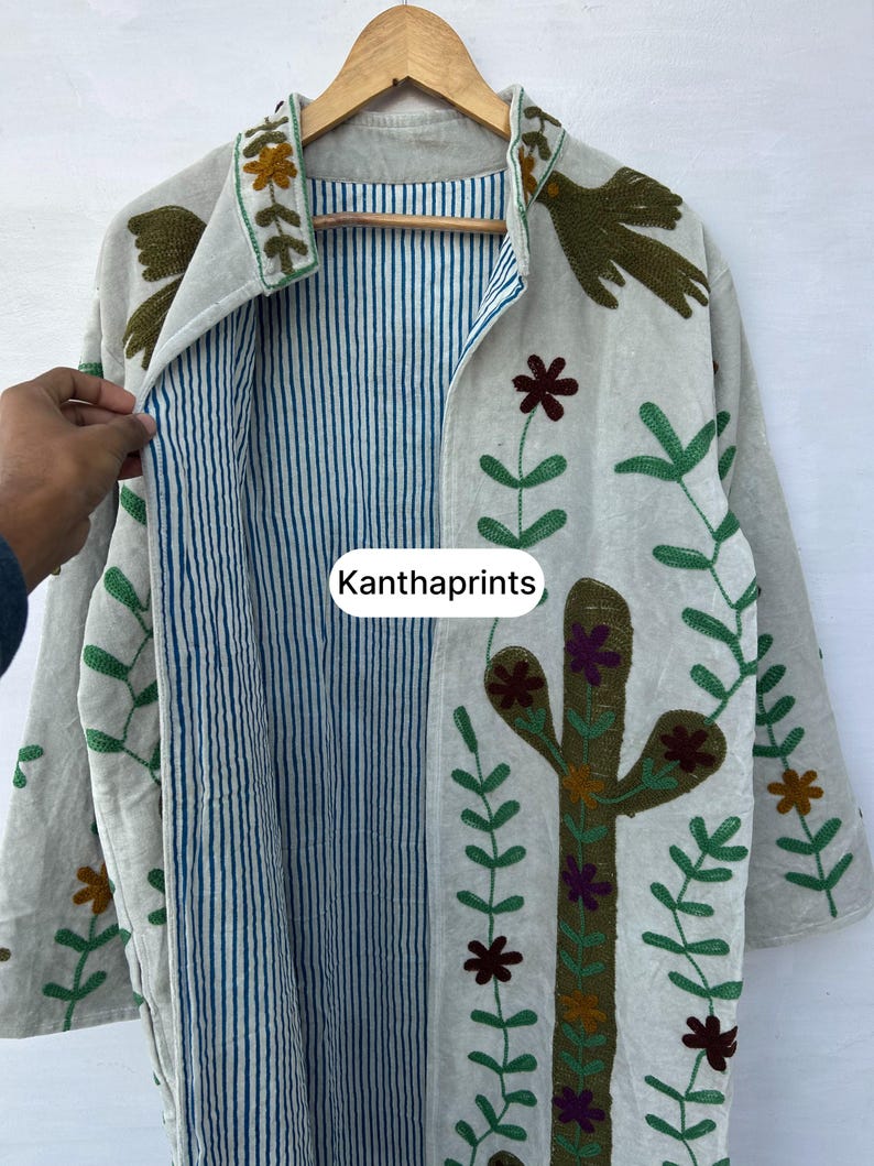May include: Beige embroidered jacket with a blue and white striped interior. The jacket features floral and cactus embroidery in green, brown, and purple. The text "Kanthaprints" is visible on the front.