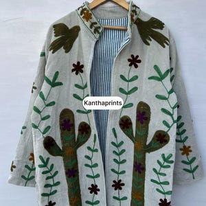 May include: A light-colored, long-sleeved jacket with embroidered cactus and floral designs. The jacket features a striped interior and is adorned with green and brown bird and flower motifs. The text "Kanthaprints" is visible on the jacket.