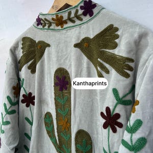 May include: A light gray jacket with embroidered details. The jacket features a cactus, birds, and floral designs in shades of green, brown, purple, and yellow. The text "Kanthaprints" is visible on the jacket.