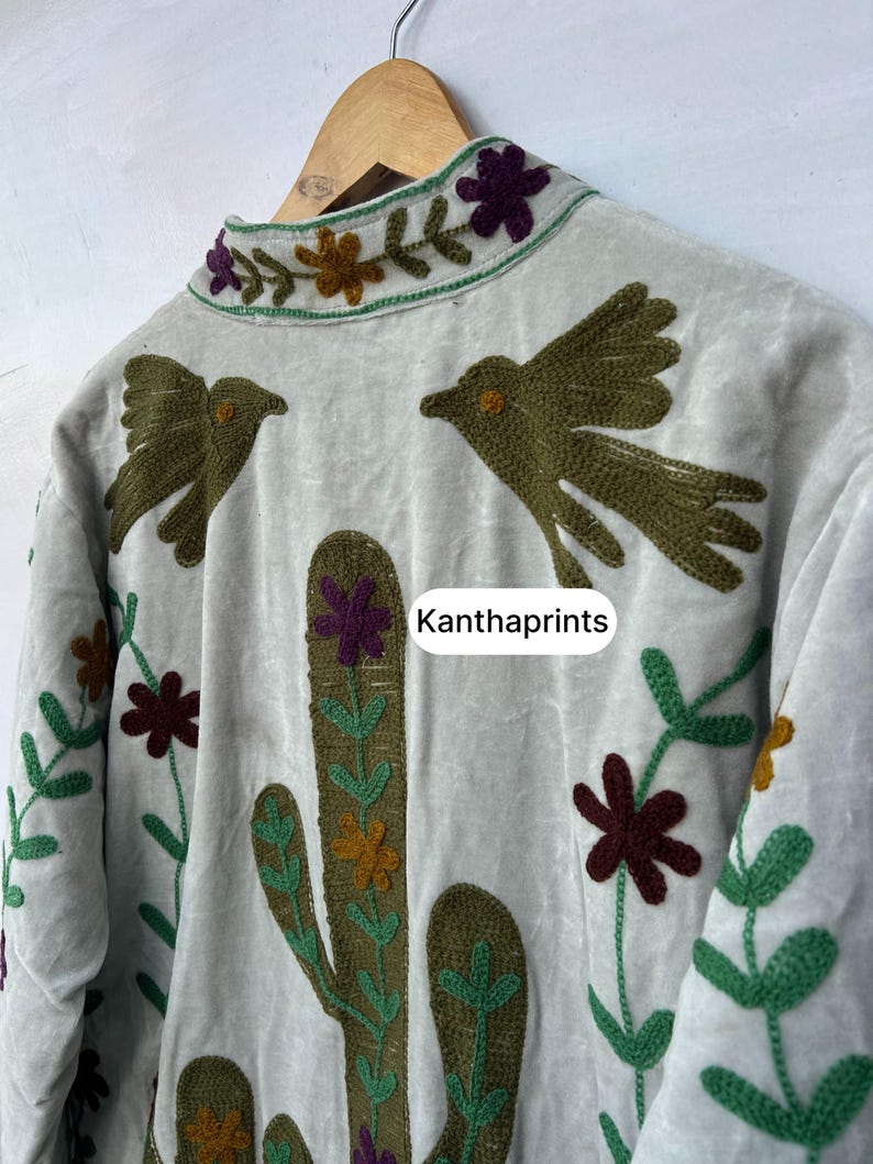 May include: A light gray jacket with embroidered details. The jacket features a cactus, birds, and floral designs in shades of green, brown, purple, and yellow. The text "Kanthaprints" is visible on the jacket.