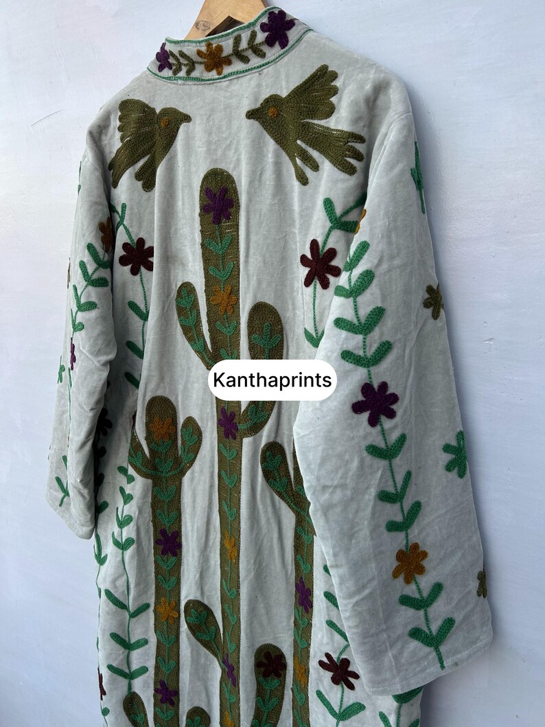May include: A light gray embroidered jacket featuring a cactus and bird design. The jacket has long sleeves and is decorated with green, brown, purple, and yellow floral and plant patterns. The text "Kanthaprints" is visible.