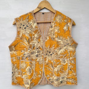 May include: A quilted vest in a mustard yellow color with a floral pattern in beige and brown. The vest has two pockets and a striped lining in brown and white. The vest is sleeveless and hangs on a wooden hanger.
