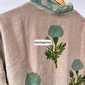 May include: A beige garment with a high collar and floral embroidery in teal. The garment features teal floral designs on the front. The text "Kanthaprints" is visible on the garment.