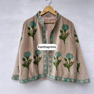 May include: Beige jacket with teal floral print and trim. The jacket is reversible, with a striped pattern on the inside. The text "Kanthaprints" is visible on the front. The jacket is hanging on a wooden hanger.