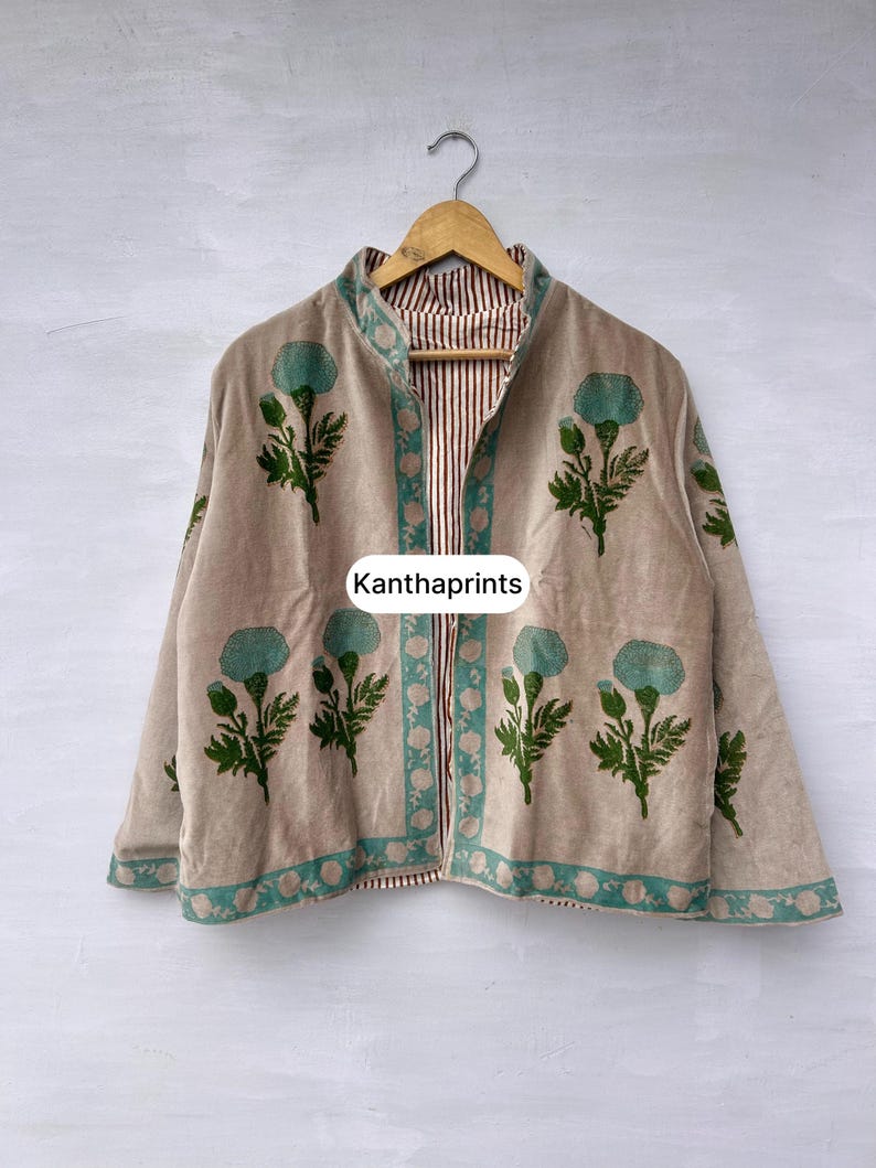 May include: Beige jacket with teal floral print and trim. The jacket is reversible, with a striped pattern on the inside. The text "Kanthaprints" is visible on the front. The jacket is hanging on a wooden hanger.