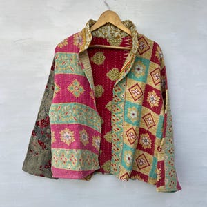 Può includere: Giacca trapuntata colorata con un design patchwork unico. La giacca presenta un mix di motivi, tra cui forme floreali e geometriche, nelle tonalità del rosa, verde acqua, oro e rosso. È appesa a una gruccia di legno su sfondo bianco.