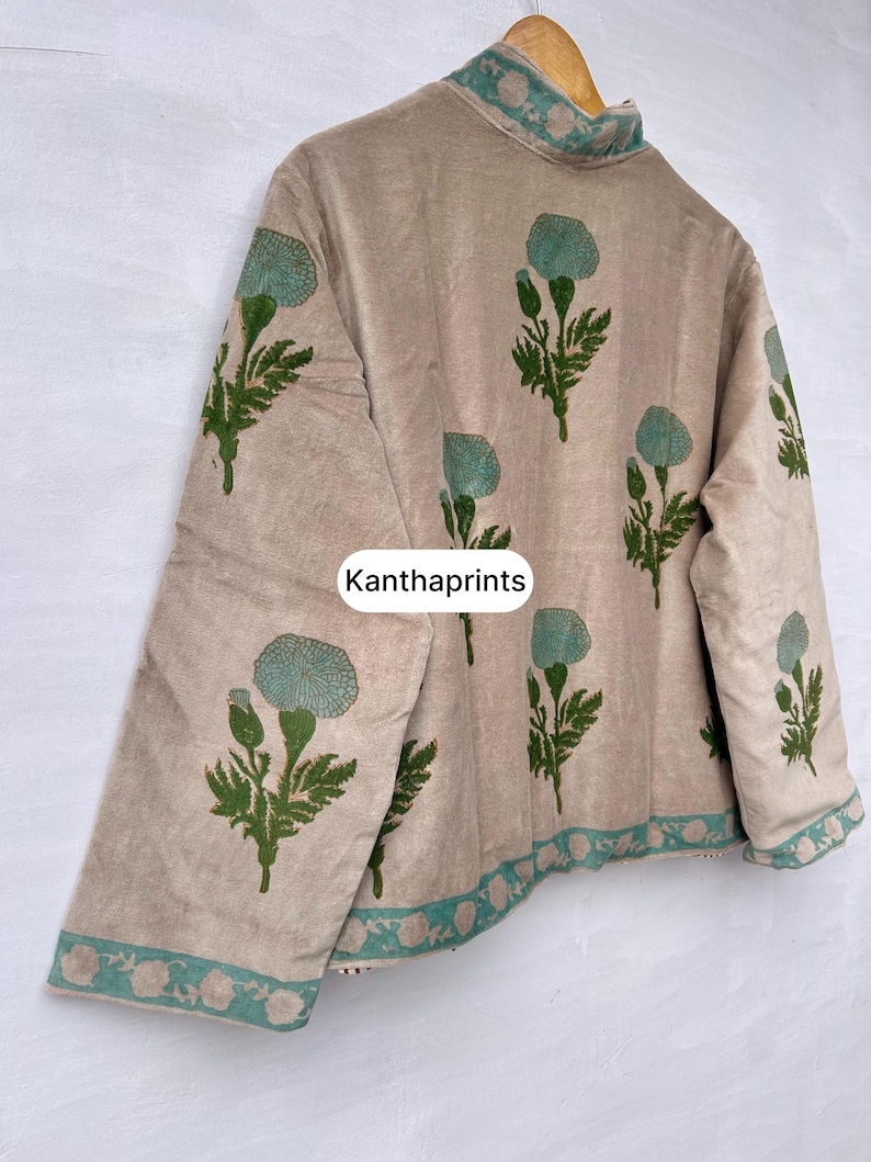 May include: Beige jacket with a floral pattern in green and teal. The jacket has a high collar and is decorated with a repeating floral design. The text "Kanthaprints" is visible on the front of the jacket.