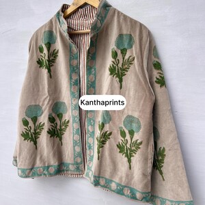 May include: Beige jacket with a floral pattern in teal and green. The jacket has a striped lining and a teal border. The text "Kanthaprints" is visible on the front. The jacket is hanging on a wooden hanger.