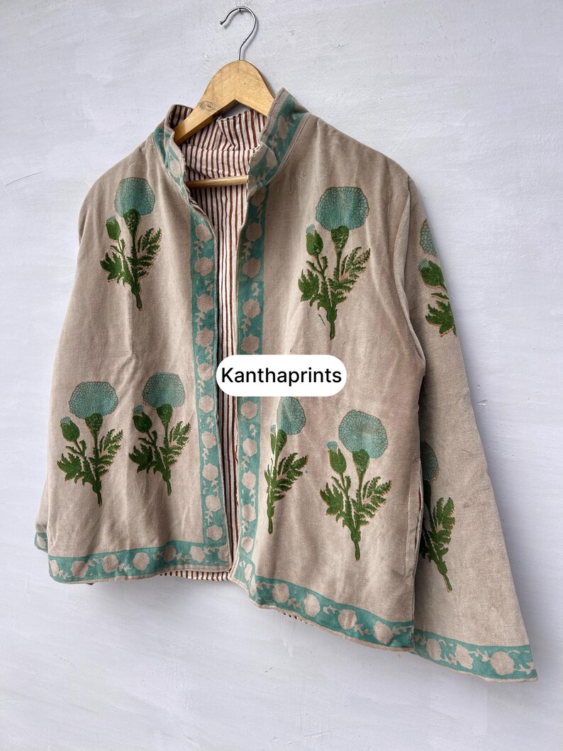 May include: Beige jacket with a floral pattern in teal and green. The jacket has a striped lining and a teal border. The text "Kanthaprints" is visible on the front. The jacket is hanging on a wooden hanger.