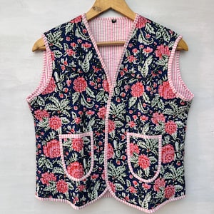 May include: A sleeveless quilted vest with a floral pattern. The vest features a navy blue background with red flowers and green leaves. The edges and pockets are trimmed with pink and white striped fabric. The vest is hanging on a wooden hanger.
