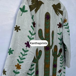 May include: A light-colored, long-sleeved jacket with embroidered details. The jacket features a cactus design, birds, and floral patterns in shades of green, brown, and purple. The text "Kanthaprints" is visible on the front.