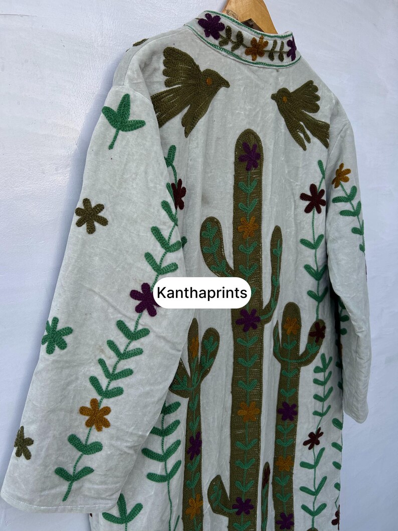 May include: A light-colored, long-sleeved jacket with embroidered details. The jacket features a cactus design, birds, and floral patterns in shades of green, brown, and purple. The text "Kanthaprints" is visible on the front.
