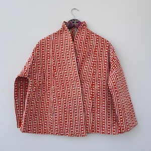 Może przedstawiać: Rdzawo-czerwona kurtka z białym wzorem w romby i pionowe paski. Kurtka ma wysoki kołnierz i wisi na srebrnym wieszaku. Kurtka jest wykonana z lekkiej tkaniny.