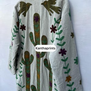 May include: A light gray embroidered jacket featuring a cactus and bird design. The jacket has long sleeves and is decorated with green, brown, purple, and orange floral and plant patterns. The text "Kanthaprints" is visible.