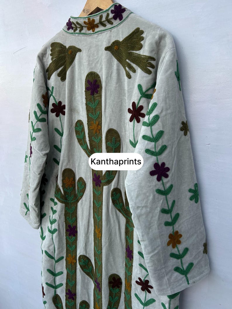 May include: A light gray embroidered jacket featuring a cactus and bird design. The jacket has long sleeves and is decorated with green, brown, purple, and orange floral and plant patterns. The text "Kanthaprints" is visible.
