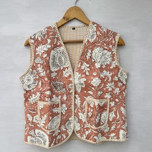May include: A quilted vest with a floral pattern in shades of coral and white. The vest has a V-neck, two front pockets, and a cream-colored trim. It hangs on a wooden hanger against a neutral background.