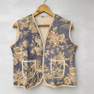 May include: A reversible quilted vest with a floral pattern in gold and gray. The vest has two front pockets and a V-neck. The interior lining is a cream and white checkered pattern. The vest is hanging on a wooden hanger.