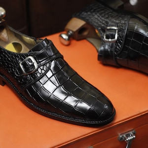 Peut inclure: Une paire de chaussures à boucle de couleur noire avec une texture en cuir gaufré façon crocodile, une boucle en métal argenté et un embauchoir en bois inséré dans chaque chaussure, posées sur une surface orange plane.