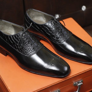 May include: A pair of black leather oxford shoes featuring a polished finish, a textured crocodile-embossed leather panel across the lace-up section, and decorative brogue detailing on the rounded toe caps with thin black laces.