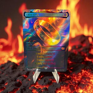 Op de afbeelding: Een holografische ruilkaart met een gouden ring met inscriptie. De kaart heeft een regenboogglans en wordt op een transparante standaard gepresenteerd tegen een vurige achtergrond. De kaart is genummerd 001/001.