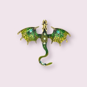 Könnte beinhalten: Eine grüne und goldene Drachenbrosche mit klaren Kristall-Akzenten. Der Drache hat ausgebreitete Flügel und einen eingerollten Schwanz.