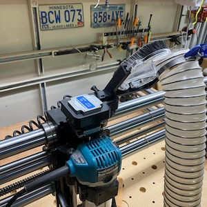 May include: A close-up of a router with a dust collection system attached. The router is blue and black and has a clear plastic dust collection tube. The dust collection system is attached to a metal frame with a spring.