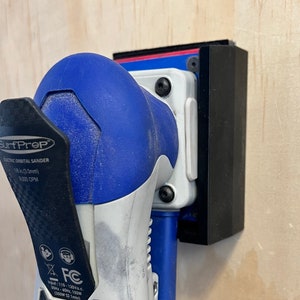 Puede incluir: Una lijadora orbital eléctrica azul y negra con un soporte de montaje negro. La lijadora tiene una etiqueta que dice "Surf Prep Electric Orbital Sander 5/8 in (15mm) 1000 OPM" y "FC" con una lista de especificaciones.