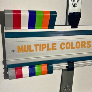Pu&ograve; includere: Un righello in metallo argentato e verde acqua con un gancio nero a ciascuna estremit&agrave;. Il righello ha un'etichetta che dice "MULTIPLE COLORS" in lettere arancioni. Il righello ha una fila di punte in plastica colorate in alto e in basso, tra cui grigio, rosso, bianco, blu, verde, arancione e nero.