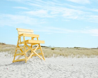May include: A yellow wooden lifeguard chair sits on a white sandy beach with tall grass in the background.