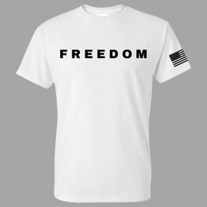 May include: White t-shirt with the word "FREEDOM" printed in large, bold, black letters across the chest. A small American flag is printed on the right sleeve. The shirt has a crew neck and short sleeves.