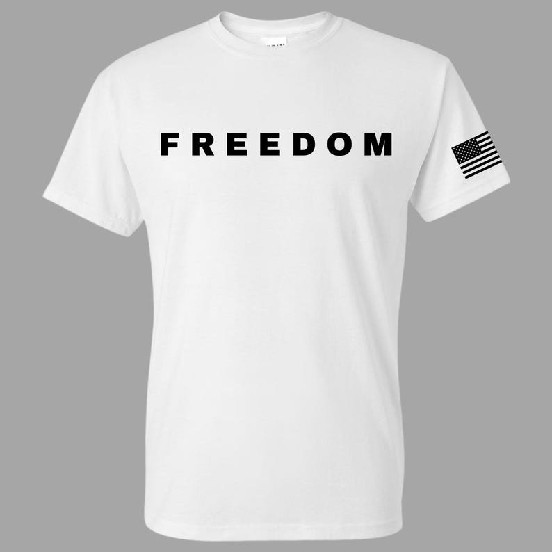 May include: White t-shirt with the word "FREEDOM" printed in large, bold, black letters across the chest. A small American flag is printed on the right sleeve. The shirt has a crew neck and short sleeves.