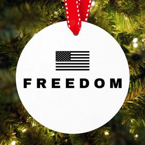 May include: White round Christmas ornament with a black and white American flag design and the word "FREEDOM" in bold black letters. The ornament hangs from a red and white ribbon, set against a backdrop of green Christmas tree branches.