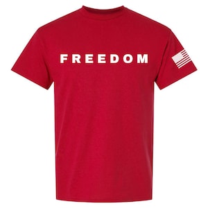 May include: A red t-shirt with the word "FREEDOM" printed in white across the chest. The American flag is printed on the right sleeve. This tee is a patriotic statement.