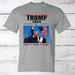 May include: Gray t-shirt with a black and white image of Donald Trump with the text "TRUMP 2024" and "FIGHT FIGHT FIGHT" printed on it.