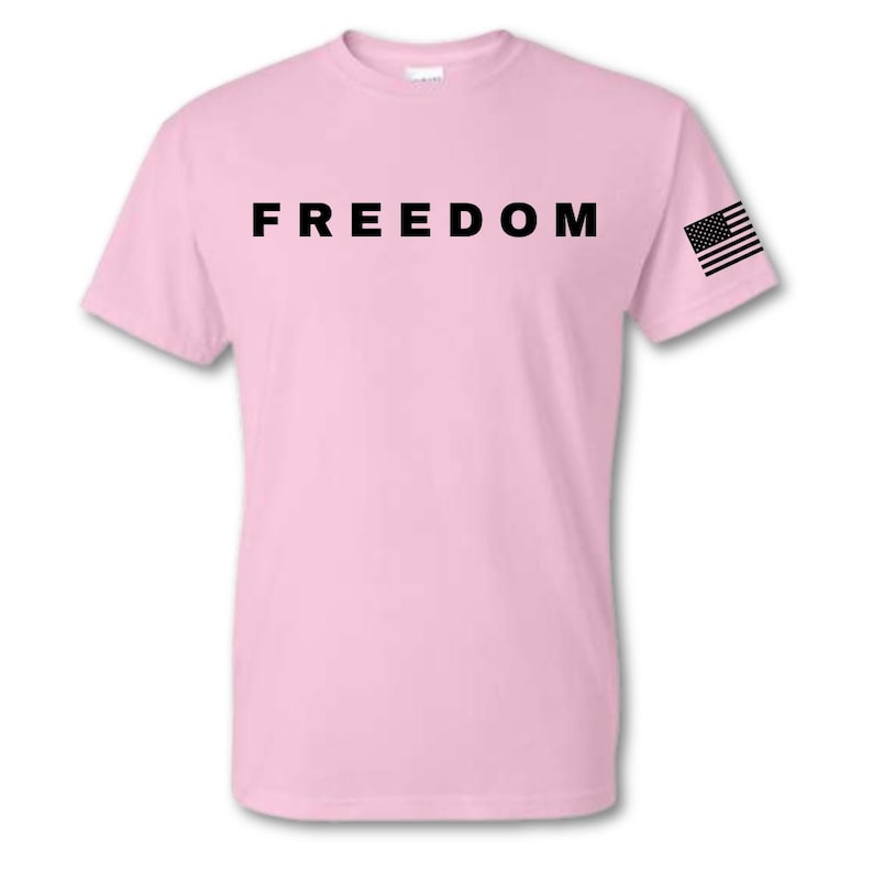 May include: A light pink t-shirt with the word "FREEDOM" printed in bold black letters across the chest. A small American flag is printed on the left sleeve. The shirt is made of a soft material.