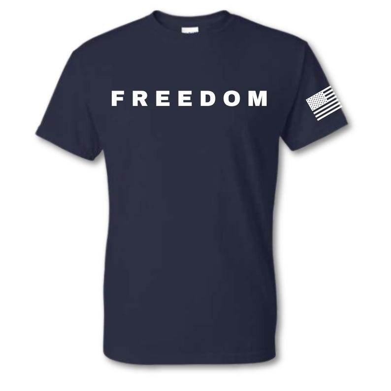 May include: Navy blue t-shirt with the word "FREEDOM" in bold white letters across the chest. A small American flag is printed on the right sleeve. The shirt is a classic crew neck style.