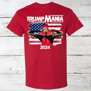 May include: Red t-shirt with a graphic of a man in a red, white, and blue bandana, holding a microphone and ripping his shirt open to reveal an American flag. The text "TRUMP MANIA" is above the graphic and "2024" is below.