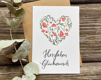 May include: A white card with a heart-shaped design made of green leaves and red flowers. The text "Herzlichen Glückwunsch" is written in black cursive lettering below the heart.