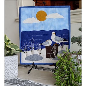 Puede incluir: Un tapiz de pared de fieltro azul y blanco con una escena de paisaje marítimo. Dos gaviotas blancas están posadas en un poste de madera con un cielo azul y nubes blancas en el fondo.