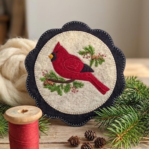 May include: A circular felt decoration featuring a red cardinal bird on a branch with green needles and small pinecones. The bird is set against a cream background, framed by a navy blue scalloped edge.