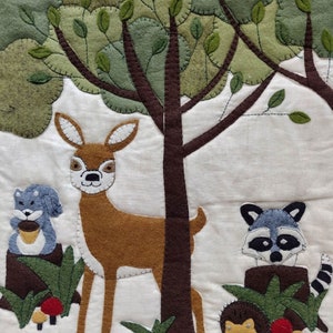 May include: A felt applique wall hanging featuring a brown deer, a gray squirrel holding an acorn, a gray raccoon, and a brown hedgehog. The animals are surrounded by green leaves and brown tree trunks. There are also red and yellow mushrooms.