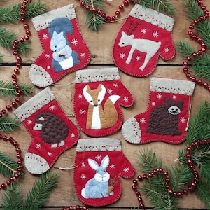 Peut inclure: Six décorations de Noël en feutre en forme de moufles, chacune représentant un animal de la forêt différent : un écureuil, un cerf, un renard, un hérisson, un ours et un lapin. Les décorations sont rouges avec des flocons de neige blancs et un poignet beige.