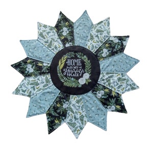 May include: A quilted, star-shaped home decor item. The design features alternating light blue and dark floral print segments. The center circle is black with the text "HOME IS MY HAPPY PLACE" surrounded by a wreath.