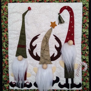 May include: Three gnomes stand in front of a reindeer with antlers. The gnome on the left wears a green hat, the gnome in the middle wears a brown hat, and the gnome on the right wears a red hat. All three gnomes have white beards and are holding a star.