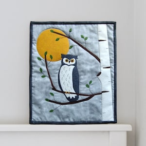 May include: A grey and white owl with large eyes sits on a branch with a yellow moon in the background. The owl is perched on a branch with green leaves. The background is a light blue with a white birch tree.