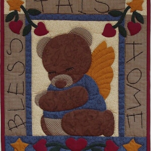 May include: A quilted wall hanging featuring a brown teddy bear wearing blue and white angel wings. The bear is holding its paws together in prayer. The background is cream with a brown and white checked border. The text "Bless This Home" is stitched in brown thread around the border.