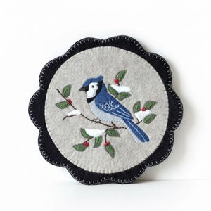 May include: Round, felt coaster with a blue jay design. The bird is perched on a branch with green leaves and red berries, set against a light gray background. The coaster has a scalloped black border.