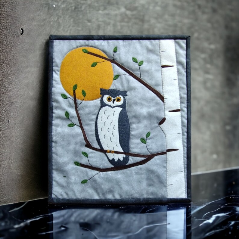 K&ouml;nnte beinhalten: Gerahmtes Textilkunstwerk mit einer Eule auf einem Ast vor einem gelben Mond. Die Eule ist haupts&auml;chlich dunkelblau und wei&szlig;, mit einem gelben Mond und gr&uuml;nen Bl&auml;ttern. Das Kunstwerk ist mit einem dunkelgrauen Rahmen versehen.