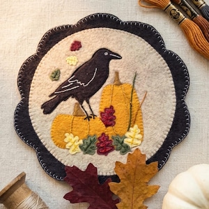 May include: A felt autumn-themed coaster with a black crow on two orange pumpkins. The coaster has a scalloped edge with a dark border and is decorated with colourful leaves.