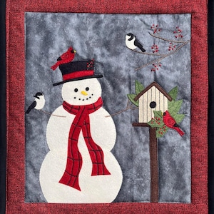 Puede incluir: Una obra de arte textil con temática invernal que presenta un muñeco de nieve con sombrero de copa negro y bufanda roja. Dos pájaros se posan en los brazos del muñeco de nieve, y un cardenal rojo está en el sombrero. También se muestra una casa para pájaros y más pájaros.